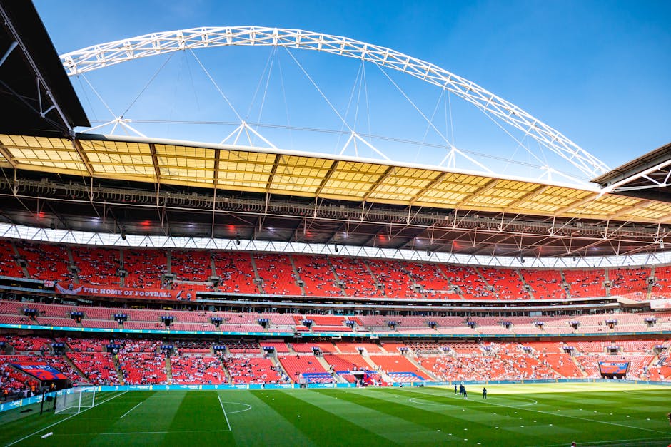 A wide view of Wembley Stadium's football pitch during daytime, featuring neatly trimmed green grass with visible mowing lines. The pitch is bordered by a clean, paved walkway and low barriers, with a