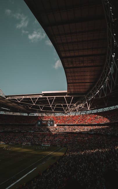 A wide view of Wembley Stadium's football pitch during daytime, featuring neatly trimmed green grass with visible mowing lines. The pitch is bordered by a clean, paved walkway and low barriers, with a
